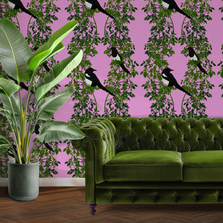 Pink wallpaper with magpies and green leaves shown with a green velvet sofa and plant